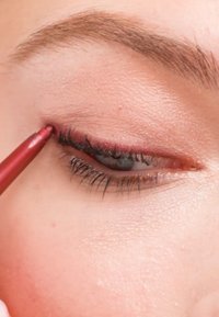 Crayon rouge appliqué sur la paupière inférieure, mettant en valeur les cils foncés et une texture de peau lisse et uniforme avec des ombres subtiles.