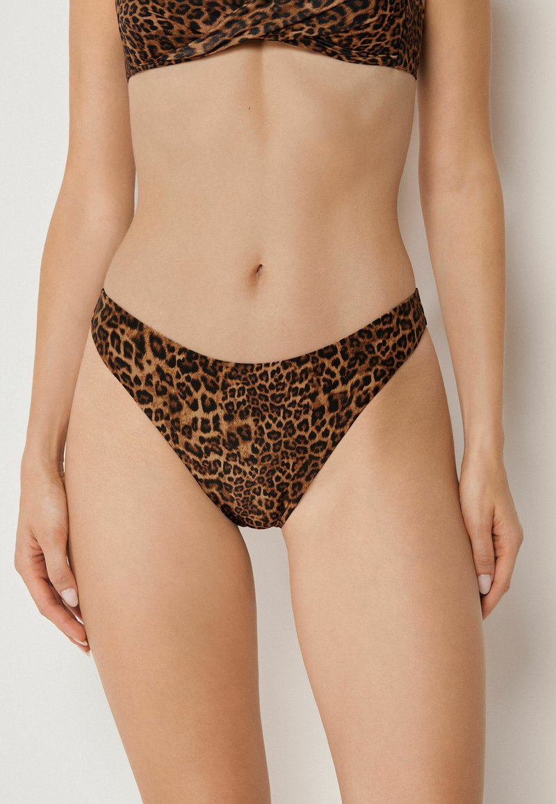 Kobieta ubrana w bikini z wzorem w cętki, stojąca z ramionami opuszczonymi wzdłuż ciała na jednolitym tle.