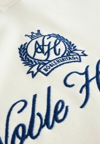 Țesătură albă cu logo brodat în albastru, prevăzut cu o coroană și o ramură de laur, textul "Patrimoniu Nobil" inscripționat într-un font cursiv, oferind textură și detaliu.