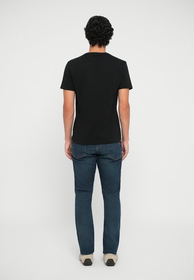 T-shirt noir à manches courtes avec un col rond, associé à un jean bleu ajusté et des chaussures de couleur claire, vu de dos.