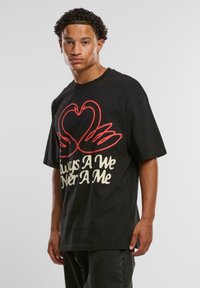 Young man with curly hair wearing a black oversized shirt featuring a red heart and hands graphic with cream colored text.