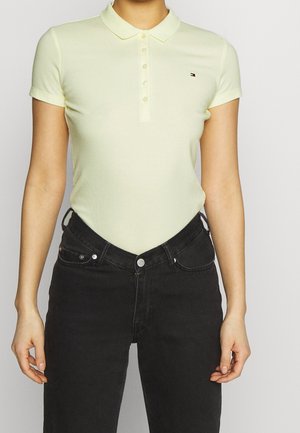 Pale yellow polo shirt with a pointed collar, short sleeves, and button placket. Worn with black high-waisted jeans. Small logo detail.