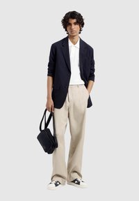 Marineblazer over een witte polo, beige broek en zwarte schoudertas. Sneakers met zwarte accenten en een witte zool. Glad textuur.