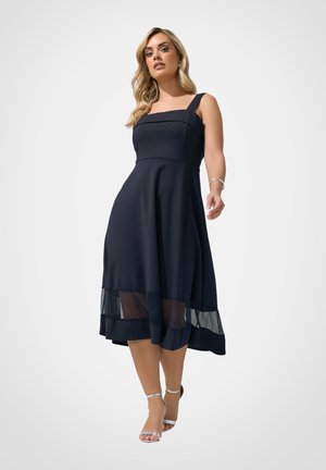 Blonde vrouw draagt een mouwloze marineblauwe midi-jurk met een vierkante halslijn en een transparant mesh paneel bij de zoom, poseert met zilveren hoge hakken sandalen.