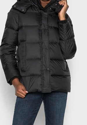 Personne portant une doudoune matelassée noire avec des boutons-pression, des poches latérales et un jean bleu foncé, tenant l'ourlet et le col de la veste.