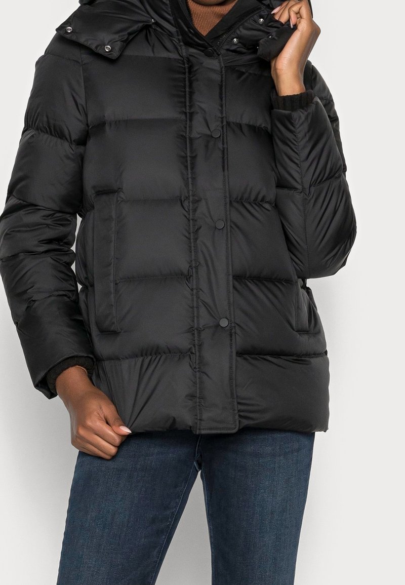 Personne portant une doudoune matelassée noire avec des boutons-pression, des poches latérales et un jean bleu foncé, tenant l'ourlet et le col de la veste.