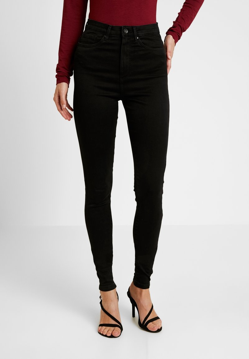 Femme portant un jean skinny taille haute noir, un haut bordeaux à manches longues et des sandales à talons hauts noires à lanières, debout sur un sol blanc.
