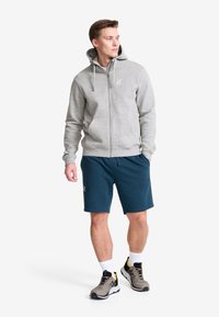 Grå zip-up hoodie med dragsnören, framfickor, i kombination med mörk teal shorts och grå sneakers med gula detaljer. Vita strumpor synliga.