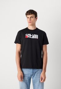 Fekete pamut póló, amelynek elején egy feltűnő fehér és piros "Calvin Klein" logó látható. Rövid ujjú, kerek nyakú, laza szabású.