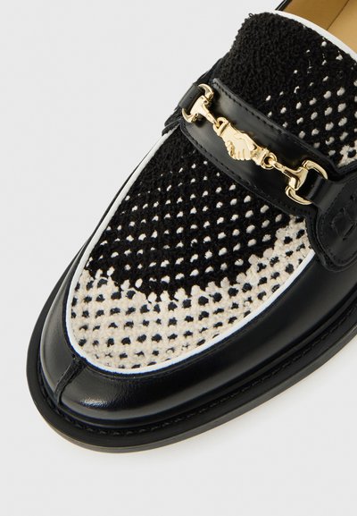Black leather loafer with black and white knitted upper and gold metal decoration shaped like two shaking hands.