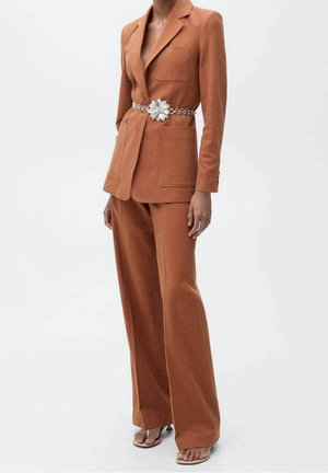 Traje naranja a medida con pantalones de pierna ancha y una chaqueta entallada. Un cinturón floral plateado acentúa la cintura. Tela suave, diseño minimalista.