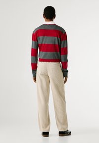 Sarkans un pelēks svītrots garajās piedurknēs krekliņš ar ribotu tekstūru. Apvienots ar bejām korduroja platmalu biksēm un melniem apaviem. Skats no aizmugures.