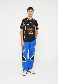 Juoda sportinė marškinė su geltonais ir baltais užrašais ant rankovių, derinami su mėlynomis kargo kelnėmis su juodais akcentais ir balta numeriu trim.