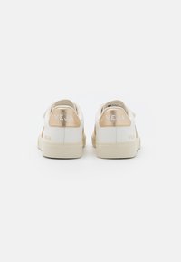 Zapatillas de cuero con un cuerpo blanco, acentos metálicos dorados en el talón y una suela beige. Logo "VEJA" exhibido en ambos talones.