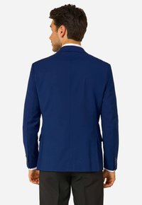 Vīrietis, kas stāv pretējā virzienā, ar piegulošu karaliskzilu bleizeri virs baltas krekla un melniem biksēm uz vienkārša fona.