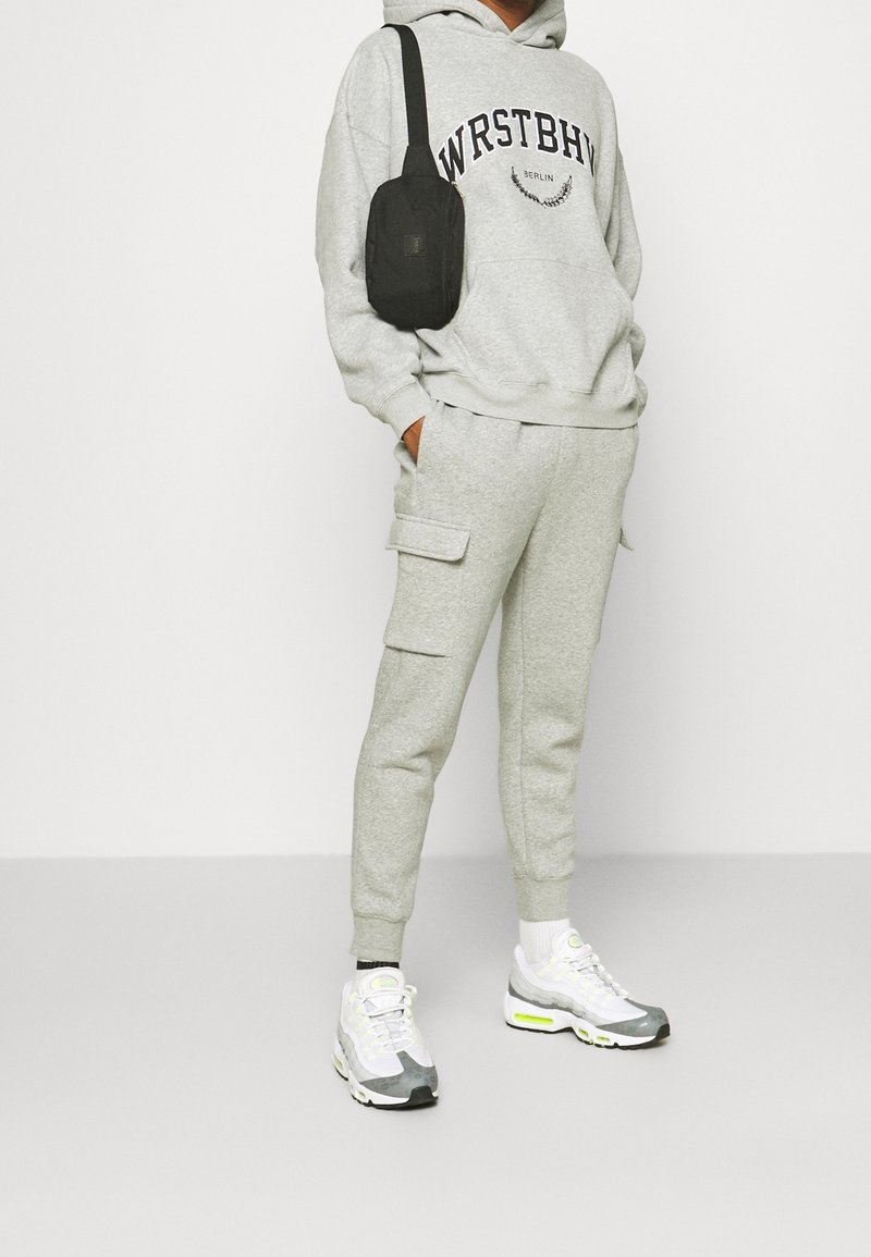 Personne portant un sweat à capuche gris clair avec le texte "WRSTBH Berlin", un pantalon cargo assorti, des baskets blanches et un petit sac à bandoulière noir.