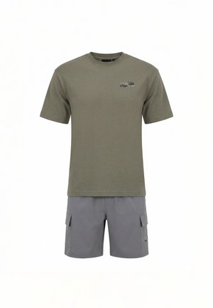 Man draagt een olijfgroen grafisch T-shirt en grijze cargoshorts, staand met de rug naar de camera op een witte achtergrond.