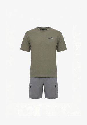 Uomo che indossa una t-shirt grafica verde oliva e pantaloncini cargo grigi, in piedi di spalle su sfondo bianco.