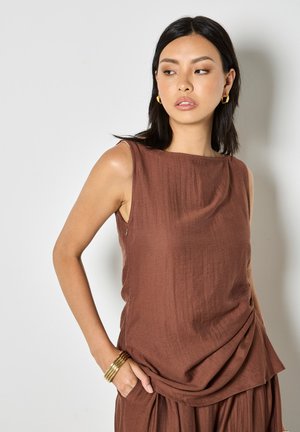 Kobieta o czarnych włosach nosi bezrękawnik w brązowym kolorze i pasującą spódnicę, złote kołowe kolczyki oraz kilka złotych bransoletek na lewej ręce.