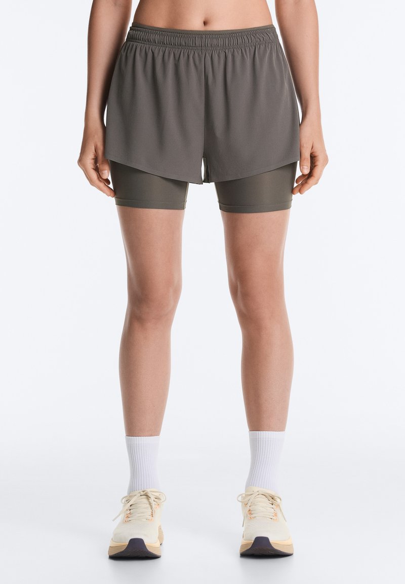 Femme portant un short de sport gris foncé à couches, des chaussettes blanches montantes et des chaussures de course beiges, debout devant un fond uni.