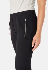 Zwarte joggingsbroek van zachte stof, met een elastische tailleband, een trekkoord en ritszakken aan de zijkanten. Soepele textuur.