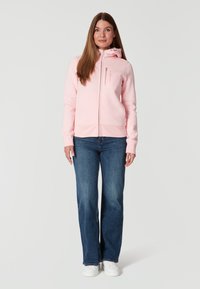 Sudadera con cremallera de color rosa claro, con capucha y bolsillos, combinada con jeans azules y zapatillas blancas. Material suave y corte relajado.