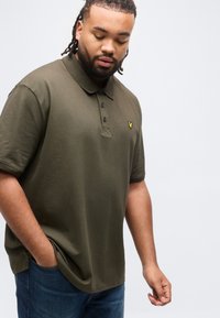 Άνδρας με πλεγμένα μαλλιά, φορώντας ένα πουκάμισο πόλο ελιάς και μπλε τζιν, με ένα χέρι στην τσέπη, μπροστά σε απλό φωτεινό φόντο.