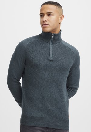 Suéter de punto de color verde azulado oscuro con un cuello de media cremallera, tejido texturizado y dobladillo y puños acanalados. Marca sutil en el lado inferior.