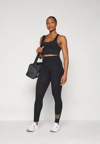 Črna atletska kratka majica s pari visokim pasom legic s rumenimi črtastimi poudarki, kombinirana z belimi supergami in črno torbo za fitnes.