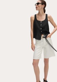 Zwarte linnen tanktop met knoopsluiting, gecombineerd met witte denim shorts met franjes aan de onderkant en een zwarte riem, voor een neutrale achtergrond.