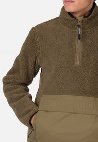 Pullover in pile di colore verde oliva con colletto alto, zip frontale a metà, ampia tasca anteriore e superficie testurizzata. Presenta dettagli con logo.