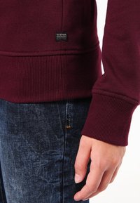 Sudadera burdeos con puños y dobladillo de canalé, presentada con jeans de mezclilla, que cuentan con una pequeña etiqueta negra de G-Star cerca de la costura inferior.