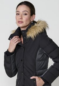 Crna puffer jakna s uklonjivim umjetnim krznom na kapuljači, s kvadratičastim uzorkom, sivim bočnim panelima i kopčama na dugmad.