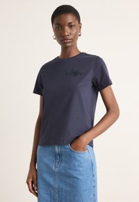 Tricou din bumbac de culoare albastru închis, cu guler rotund și mâneci scurte; prezintă un logo brodat negru pe piept. Asortat cu o fustă din denim.