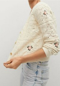 Sweter kardigan w kolorze kremowym z teksturowanym wzorem, haftowanymi akcentami kwiatowymi i drewnianymi guzikami. Posiada luźny krój i rękawy o długości trzech czwartych.