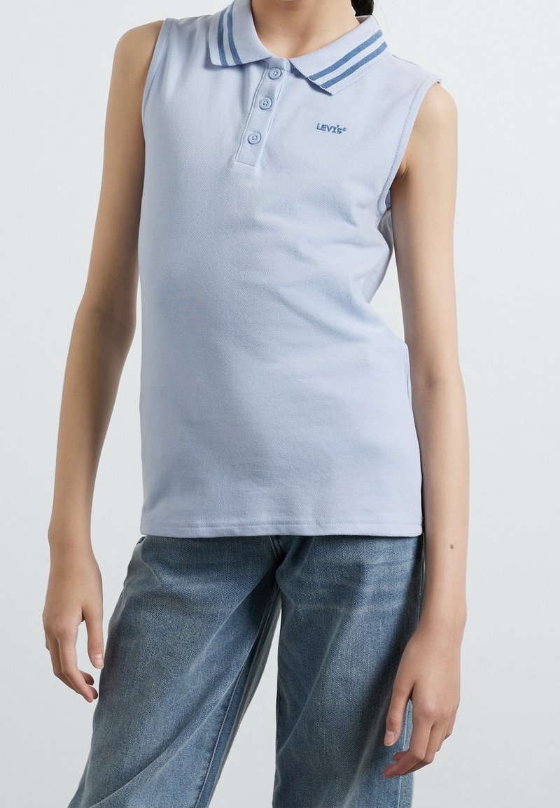 Enfant portant un polo sans manches bleu clair avec col et trois boutons, associé à un jean bleu ample sur fond clair.