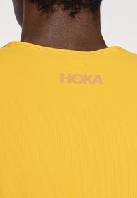 Yellow athletic shirt made of textured fabric, featuring a ribbed neckline and a subtle, light-colored logo on the back.