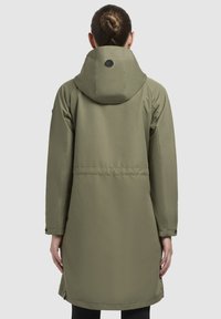 Vihreä vedenpitävä parka, jossa on huppu ja logo takana. Takissa on kireä vyötärö ja sivureiät liikkuvuuden lisäämiseksi.