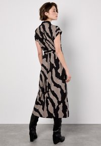 Femme portant une robe midi sans manches à motif abstrait noir et taupe, avec une ceinture, associée à des bottes noires à talons hauts jusqu'au genou, se tenant de profil.