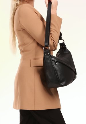 Person wearing tan blazer and black pants holds a black leather shoulder bag with a wide strap against a light background.