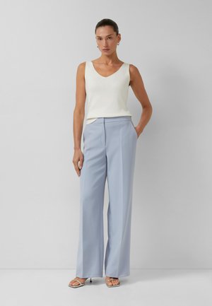 Femme debout contre un mur gris portant un haut blanc sans manches, un pantalon large bleu clair et des sandales à talons ouvertes.