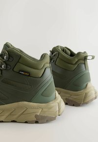 Zielone buty trekkingowe z Cordury, z teksturowaną cholewką, wzmocnionym zapiętkiem i masywną beżową podeszwą z wzórkiem bieżnika.