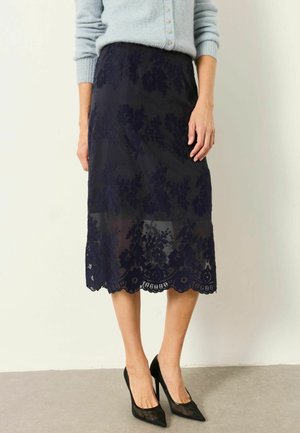 Kvinde iført en navyblå blomsterblonde midi-nederdel kombineret med sorte spidse hæle og en lyseblå knaptrøje.