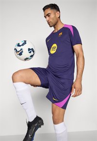 Nike Barcelona football kit in dark purple with pink accents. Player balanced on one leg, wearing black cleats and white socks. Football in mid-air.