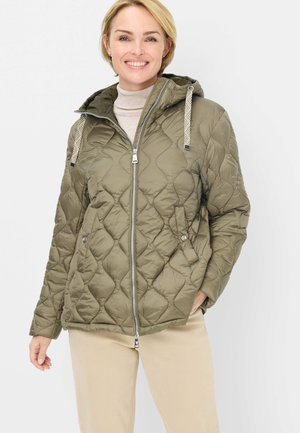 Lächelnde Frau, die eine olivgrüne gesteppte Jacke mit Kapuze und beige Hose trägt, vor einem einfachen Hintergrund stehend.