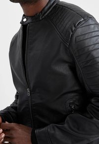 Veste en cuir noire avec fermeture éclair à l'avant, épaules matelassées et texture côtelée sur les manches. Finition lisse avec une coupe ajustée.
