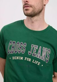 Zielona bawełniana koszulka z okrągłym dekoltem i krótkimi rękawami, z napisem "CROSS JEANS DENIM FOR LIFE" w kolorze granatowym i kremowym.