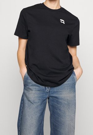 Person wearing a black T-shirt with a small white logo near the shoulder and blue faded wide-leg jeans, hands in pockets.