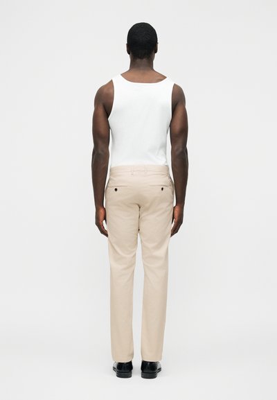 Άνδρας που στέκεται με την πλάτη, φοράει λευκή φανέλα, μπεζ παντελόνια και μαύρα παπούτσια πάνω σε απλό ανοιχτό φόντο.