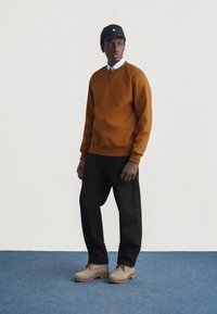 Άνδρας που φορά καφέ φούτερ, ανοιχτό μπλε πουκάμισο με γιακά, μαύρο παντελόνι, μπεζ μποτάκια και μαύρο καπέλο, στέκεται πάνω σε μπλε χαλί μπροστά σε λευκό τοίχο.